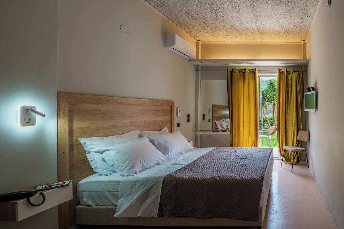 A hotel room with a bed, bedside table, lamp, and a view of the outdoors.