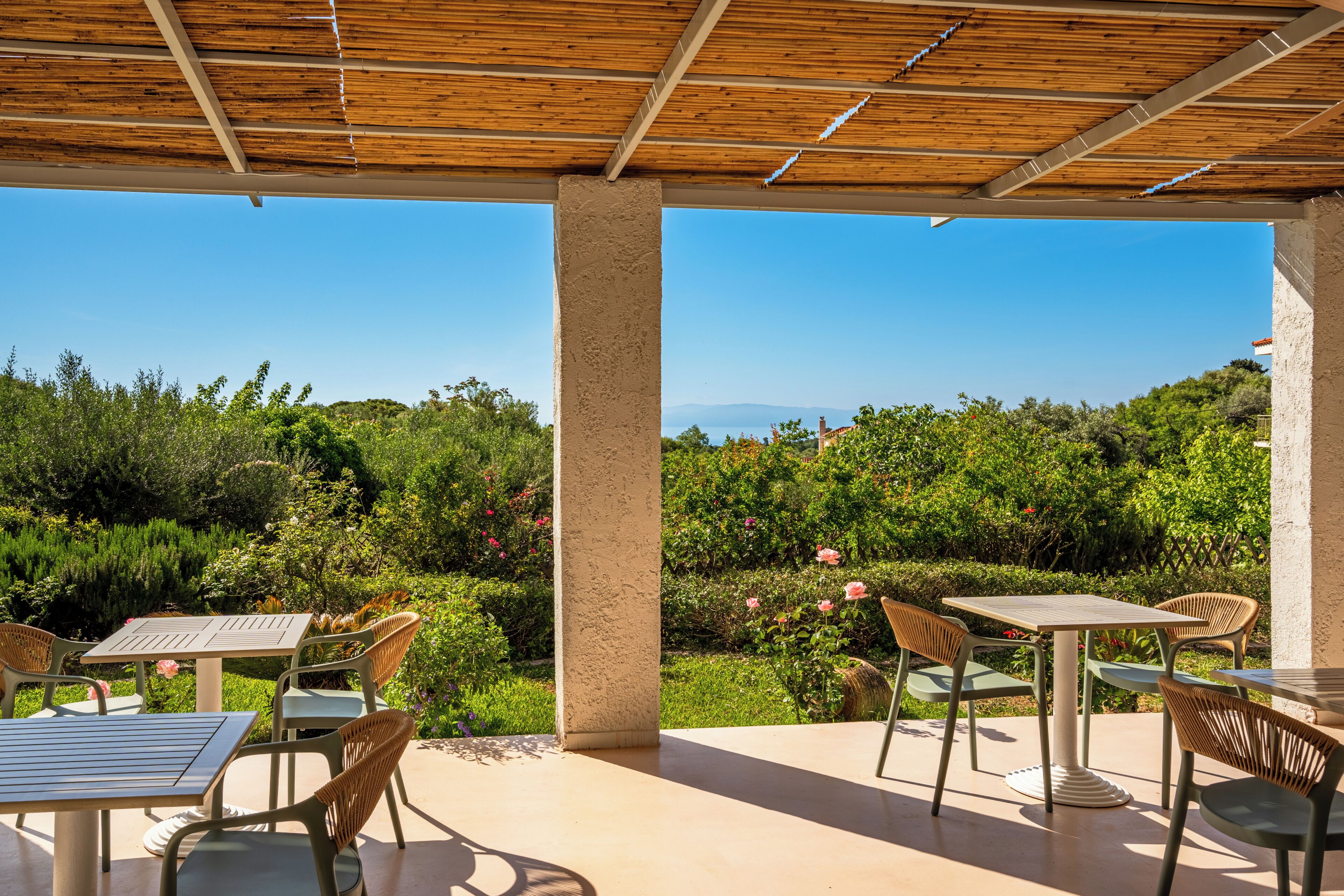 An outdoor patio with tables and chairs, a wooden ceiling, and a view of greenery and distant hills.