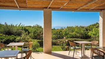 An outdoor patio with tables and chairs, a wooden ceiling, and a view of greenery and distant hills.