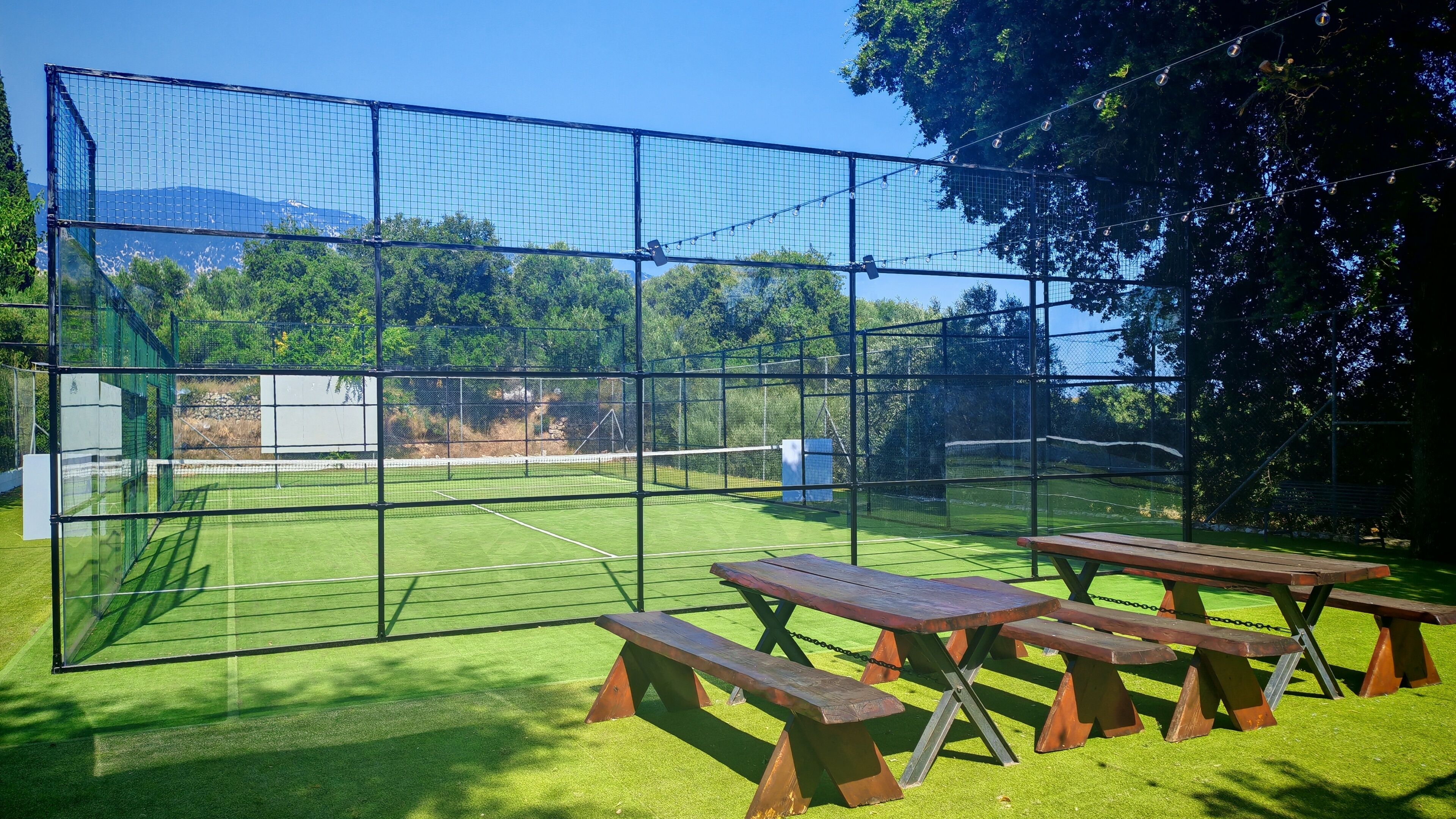 A tennis court with a net, surrounded by a fence, and picnic tables in the foreground.