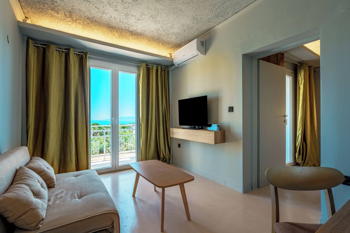 A modern living room with a sofa, a small table, a TV on a wooden shelf, and a balcony with curtains.