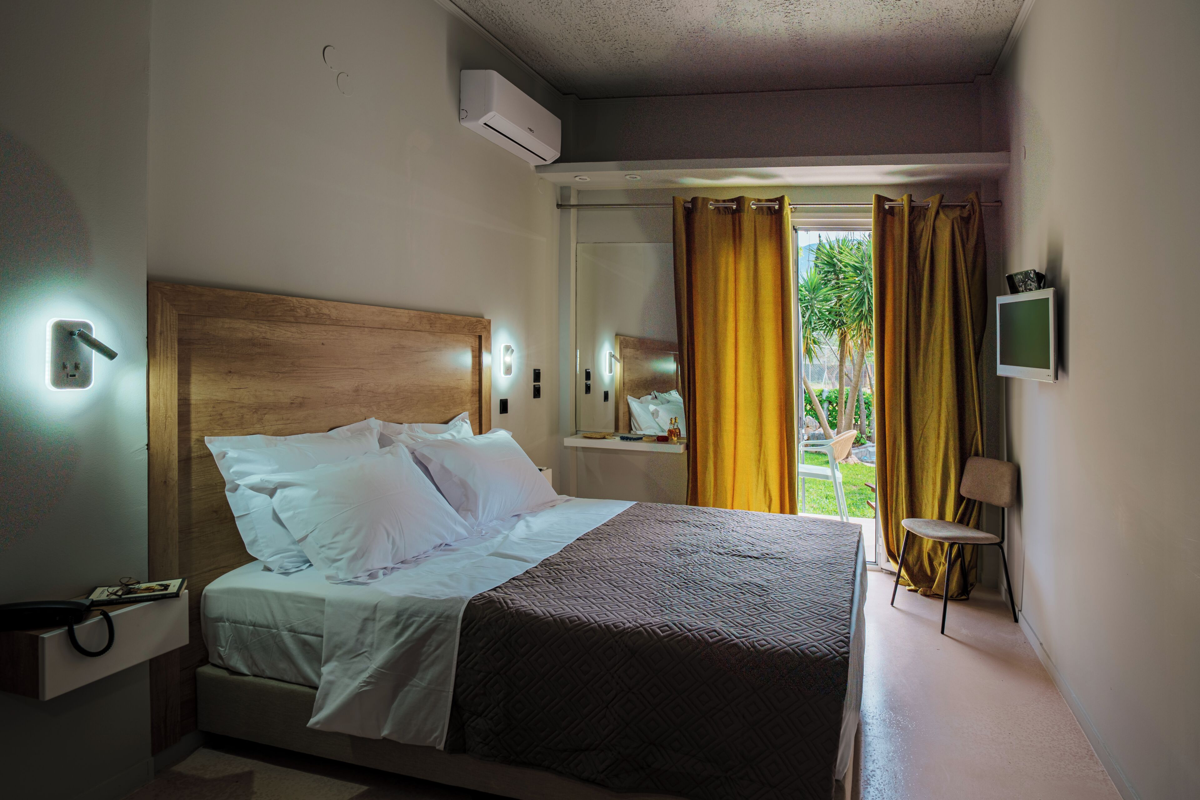 Habitación de hotel con cama, mesita de noche, televisor montado en la pared y ventana con cortinas.
