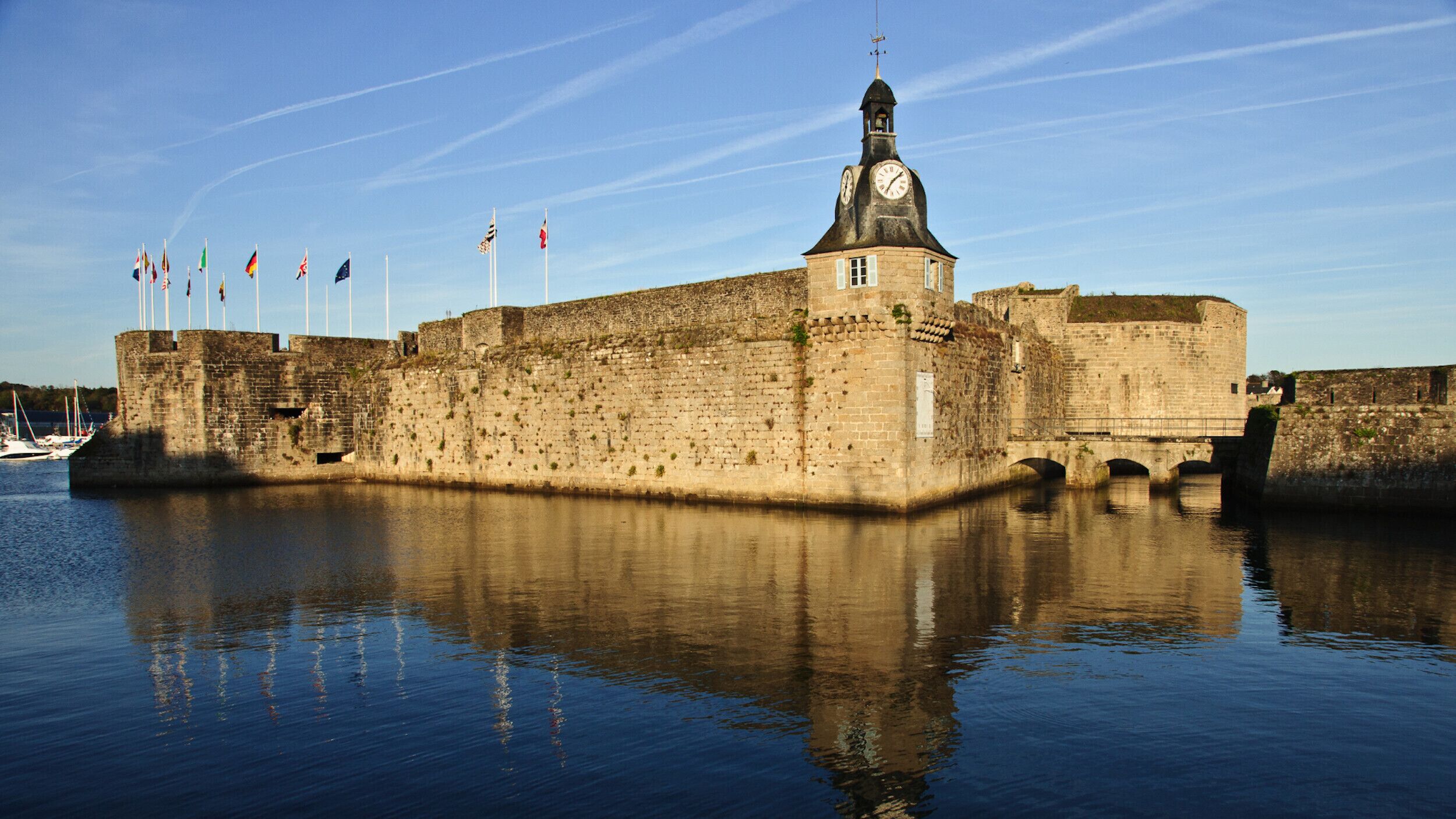 A historic stone fortress with a clock tower, surrounded by calm water and flags.