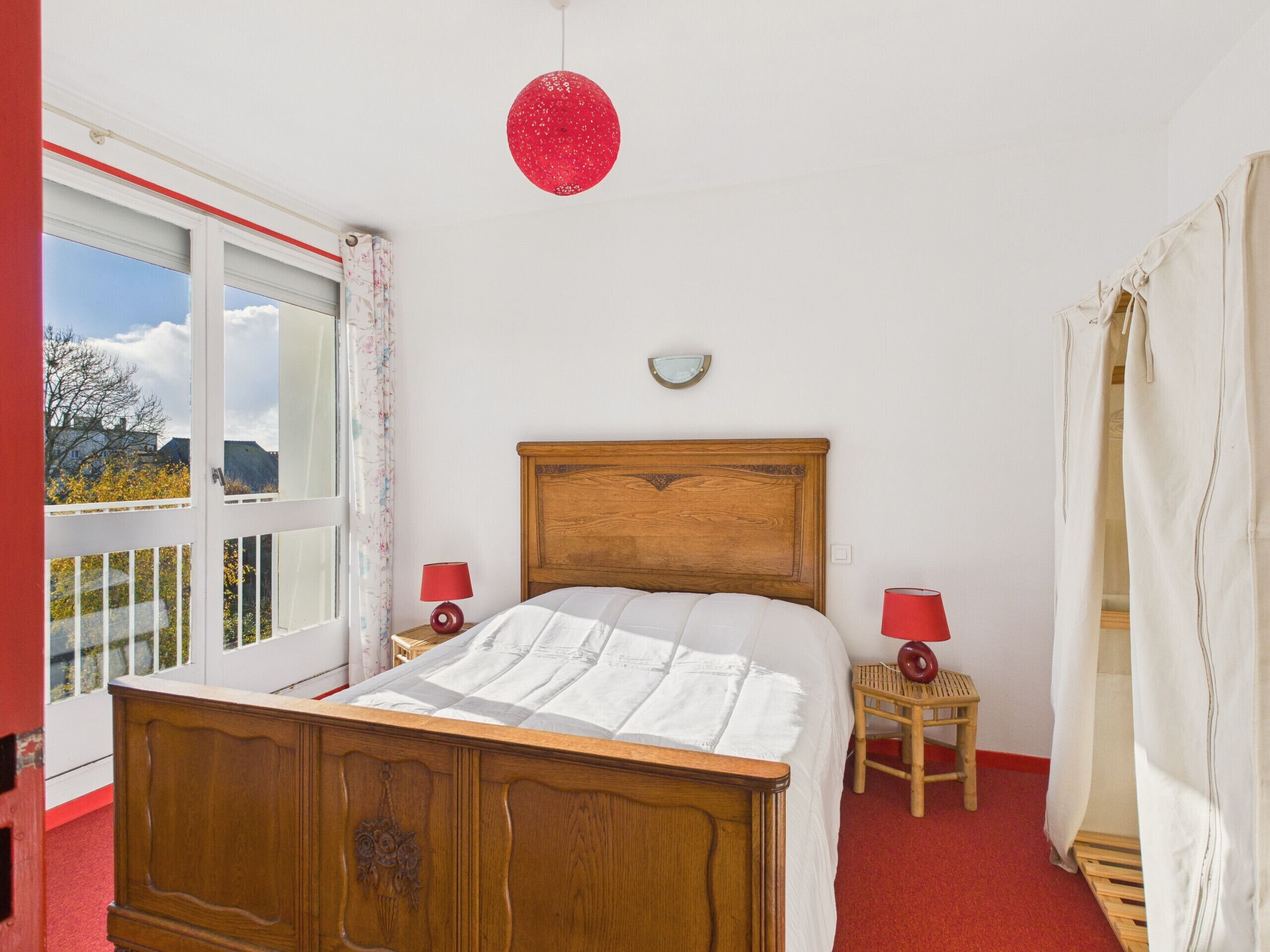 Un dormitorio con cama de madera, dos mesitas de noche con lámparas, un ventanal con cortinas y una lámpara colgante roja.