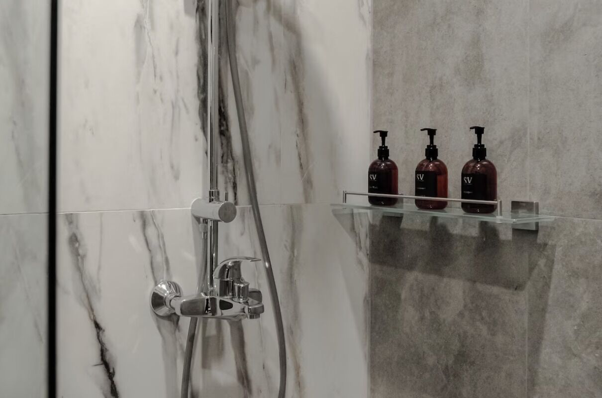 Ein Badezimmer mit Marmorwand, einer Duschkabine inklusive Handbrause und drei Shampooflaschen auf einem Regal.