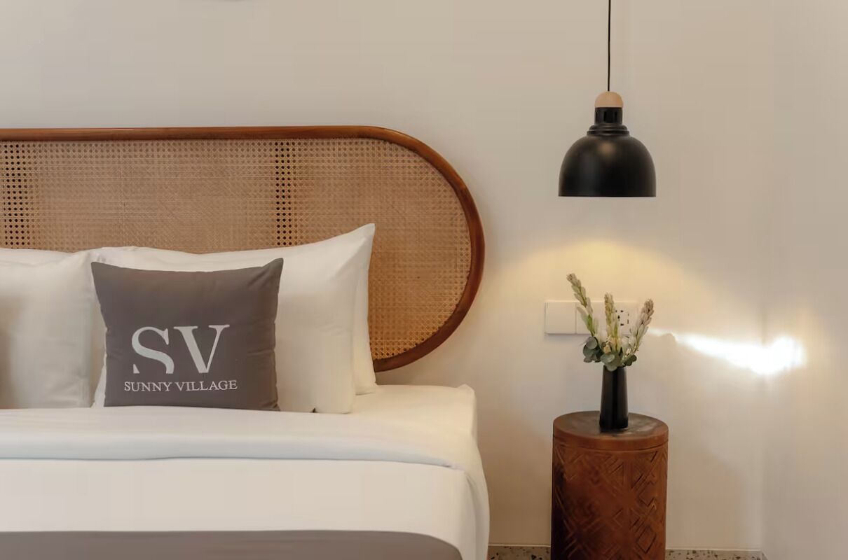 Una cama bien tendida con sábanas blancas y un cojín con el texto "SV Sunny Village", una mesita de noche redonda de madera con un jarrón de flores, y una lámpara colgante negra.