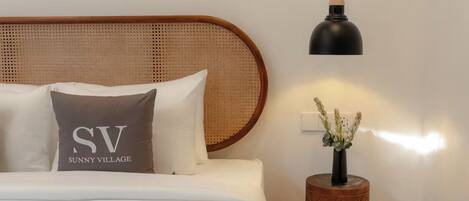 Una cama bien tendida con sábanas blancas y un cojín con el texto "SV Sunny Village", una mesita de noche redonda de madera con un jarrón de flores, y una lámpara colgante negra.