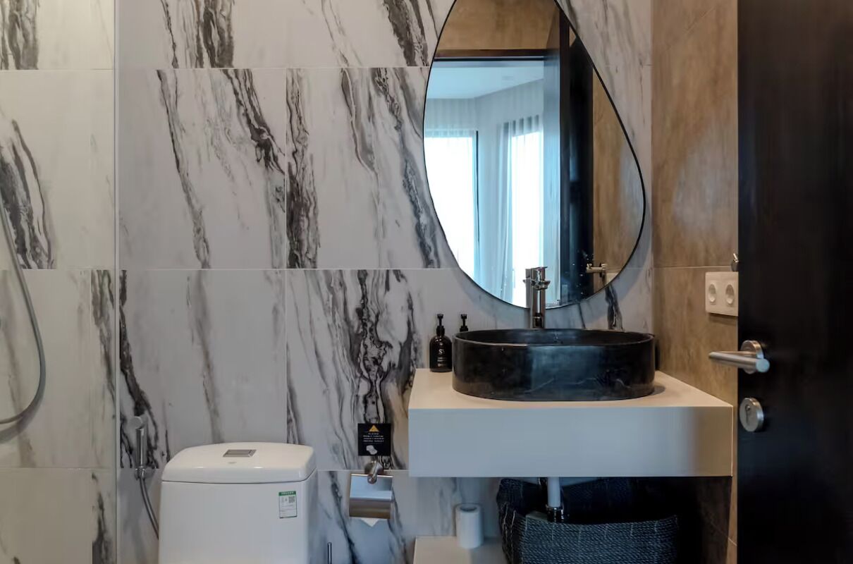 A bathroom with a marble wall, a round sink with a black basin, a toilet, and a mirror with a wooden frame.