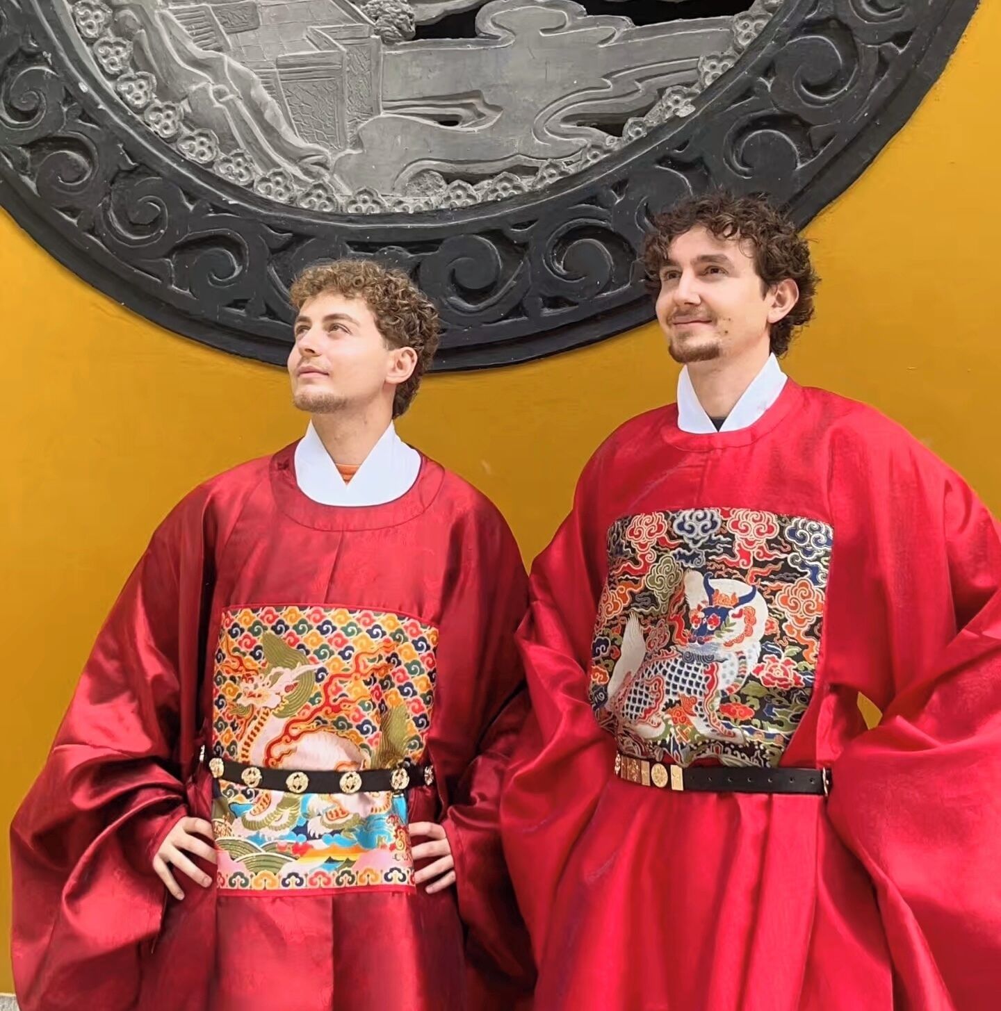 Two individuals in red robes with intricate designs, standing in front of a yellow wall with a circular emblem.