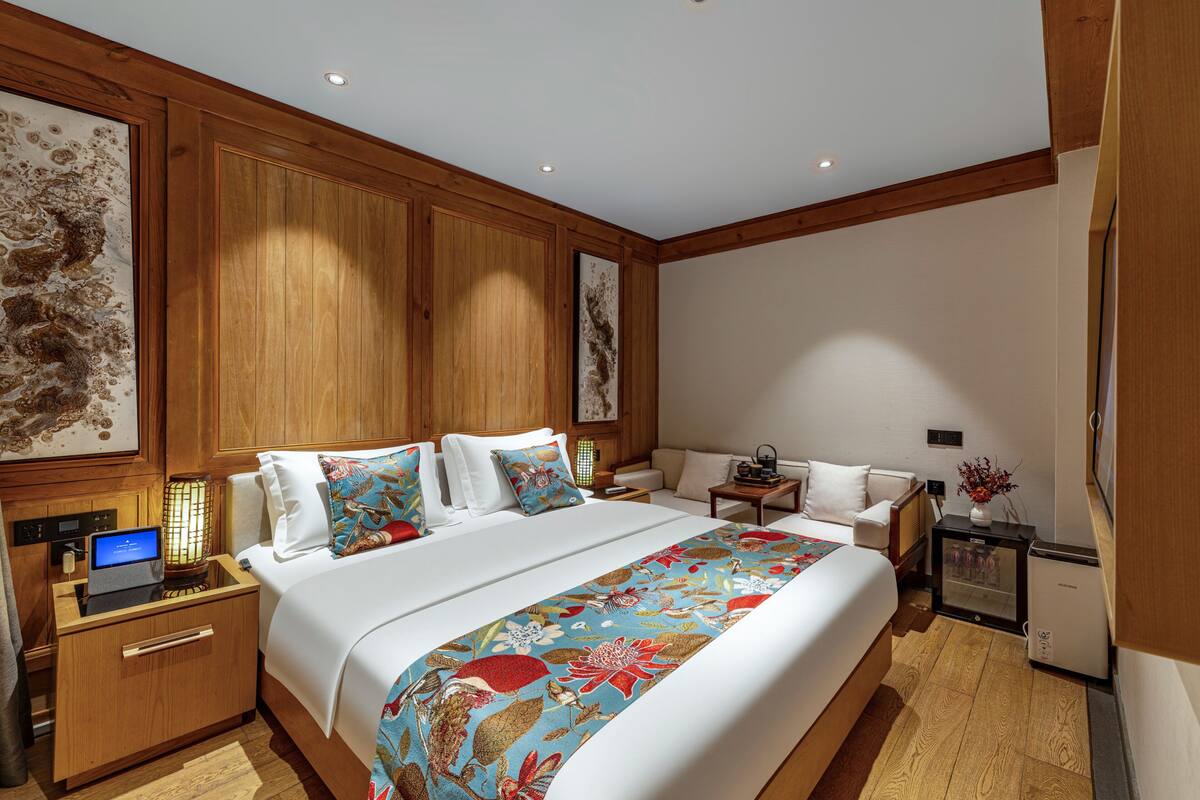 A hotel room with a large bed, a desk with a laptop, a small table with a vase, and a refrigerator.