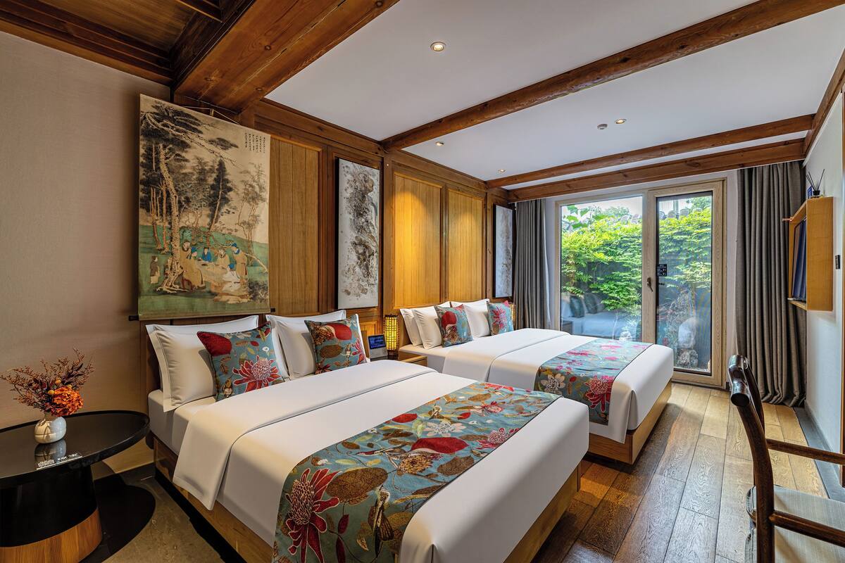 A hotel room with two beds, a large painting on the wall, and a view of greenery through the window.