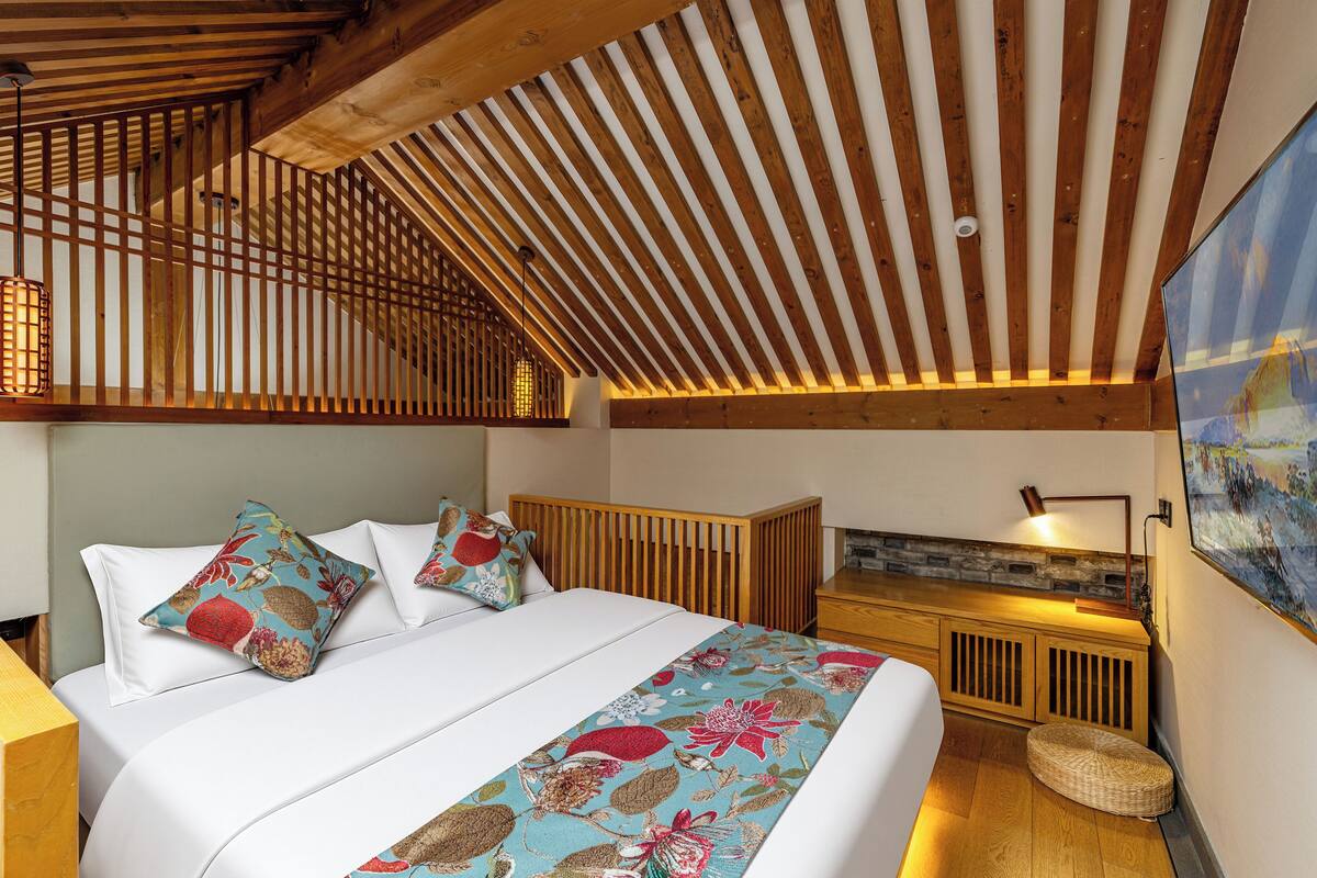 A bedroom with a sloped ceiling, a bed with floral bedding, a bedside lamp, and a painting on the wall.