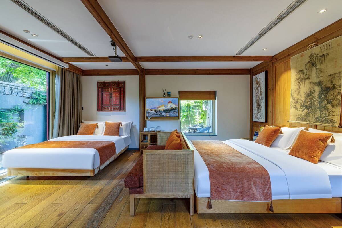 A hotel room with two beds, a view of greenery outside, and a traditional artwork on the wall.