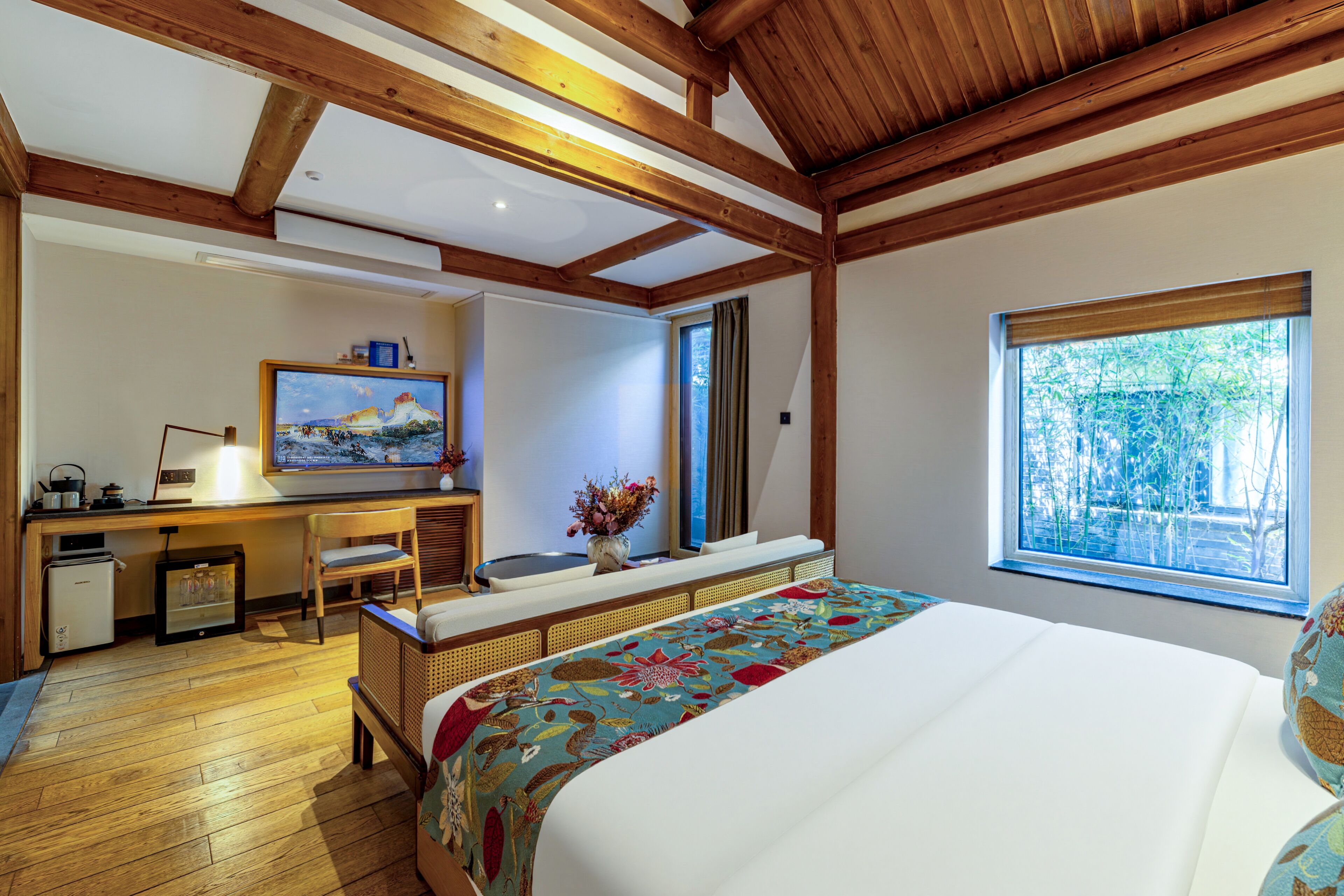 A bedroom with a bed, a desk, a chair, a small fridge, and a window with a view of trees.
