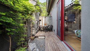 Um pátio com mesa e cadeiras, uma pequena área de jardim com plantas e um elemento decorativo em rocha, e uma porta de vidro que dá para um espaço interno.