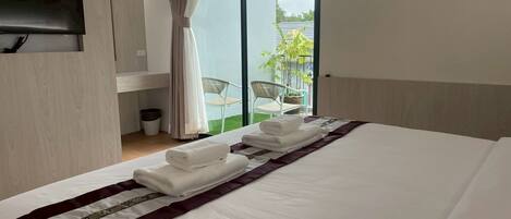 A hotel room with a large bed, a flat-screen TV mounted on the wall, and a balcony with chairs and a potted plant.