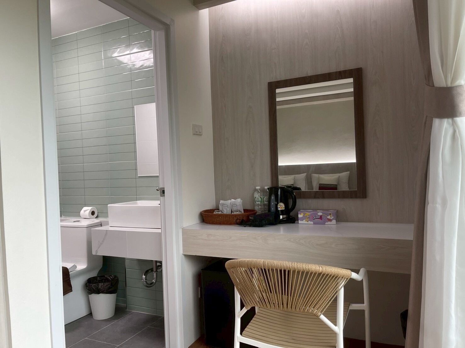 Un baño con lavamanos, inodoro y zona de ducha. Hay una silla y una mesita con artículos de aseo.