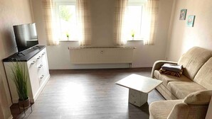 Una sala de estar con sofá, mesa de centro, televisor sobre un mueble, radiador y ventana con cortinas.