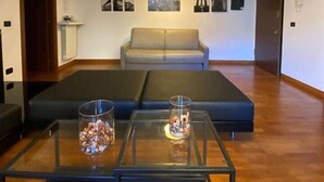Un salon moderne avec un canapé, deux tables en verre ornées d’objets décoratifs, un luminaire suspendu et des tableaux encadrés accrochés au mur.