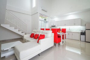 A modern living room with a white sofa, red cushions, and a dining area with red chairs. A staircase is visible on the left, and a kitchen with white cabinets is in the background. - Luxury minimalist villa The Toy (Kerobokan)