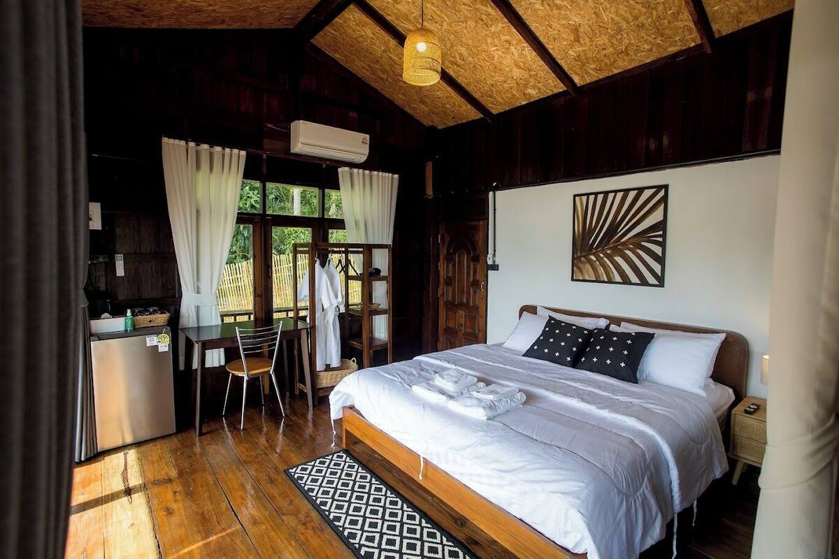 A bedroom with a wooden ceiling, a bed with white linens, a bedside table, a nightstand, and a window with curtains.