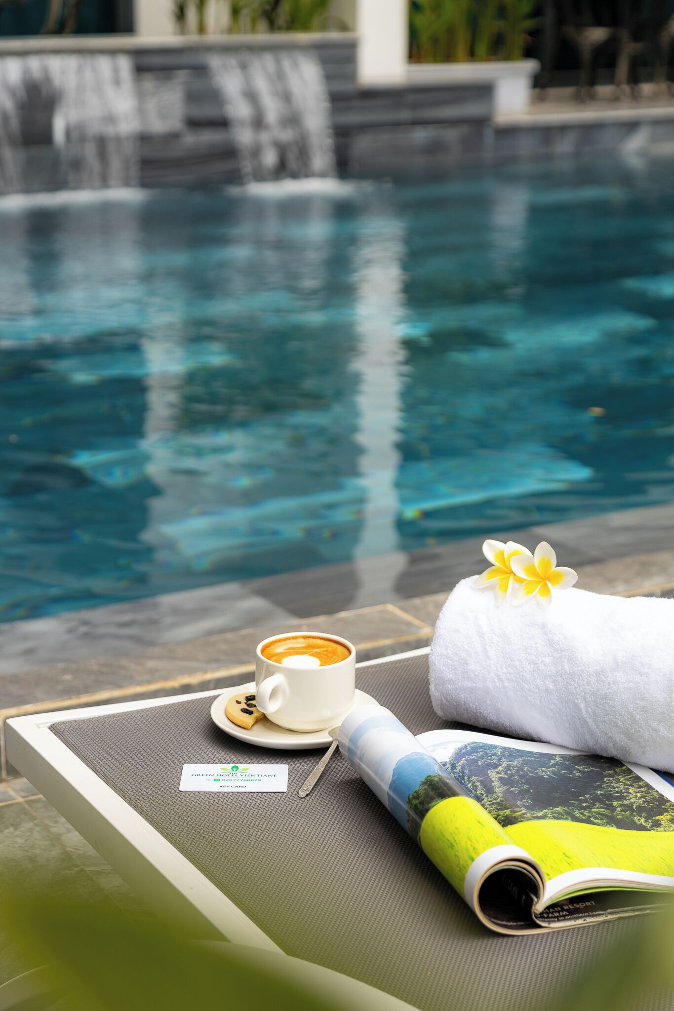 Entorn de piscina amb una tovallola blanca, una tassa de cafè i una revista damunt d'una taula.