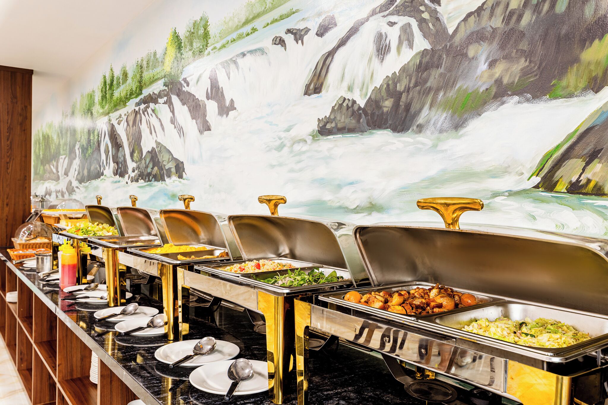 A buffet with various food options, a waterfall mural on the wall, and a wooden paneling design.
