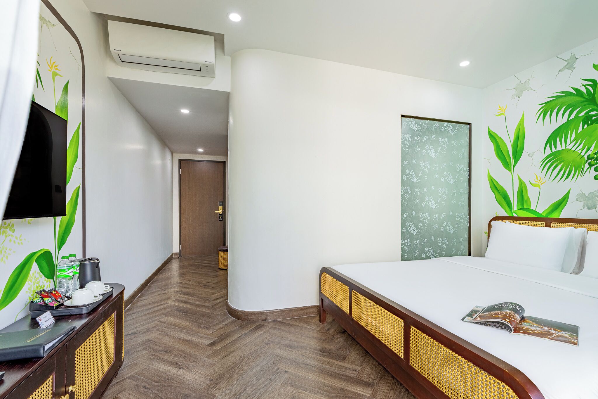 Une chambre d’hôtel comprenant un lit, une télévision, un bureau avec un plateau de petit-déjeuner, et un mur orné d’une fresque aux motifs tropicaux.