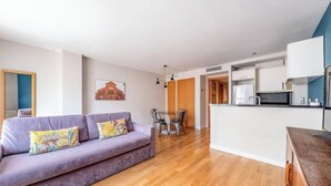 A modern living room with a purple sofa, wooden flooring, and a kitchen area with white cabinets. - 1 bedroom apartment with parking in Eixample (Barcelona)