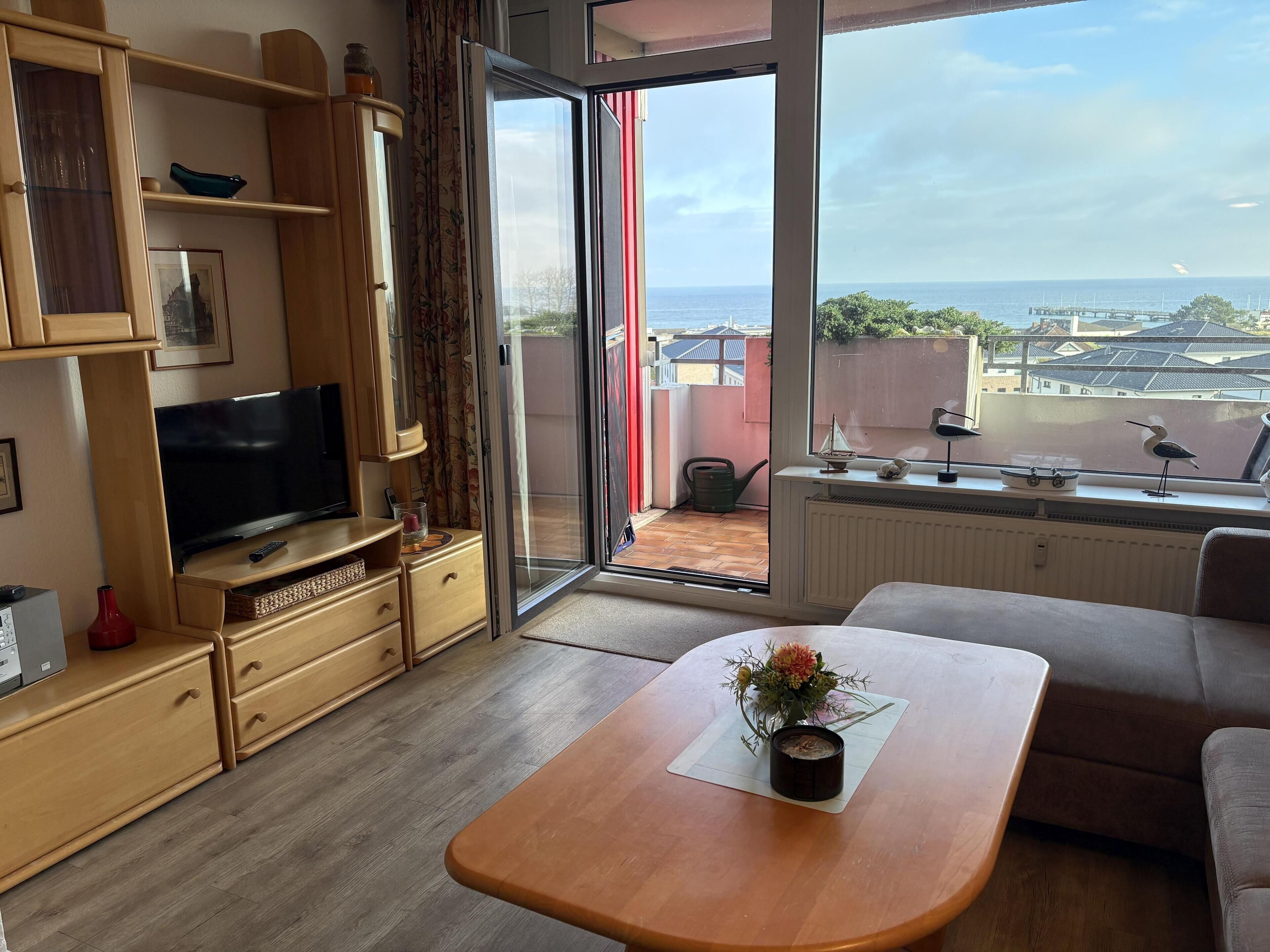 Un soggiorno moderno con televisione, mobili in legno, un balcone vista mare e un tavolo da pranzo con un vaso di fiori.