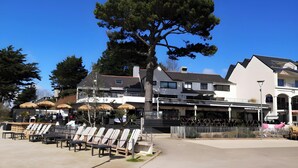 A building with a modern architectural style, outdoor seating, and a tree in front. - La Perle de l'Océan – Pool and Terrace (Guidel)