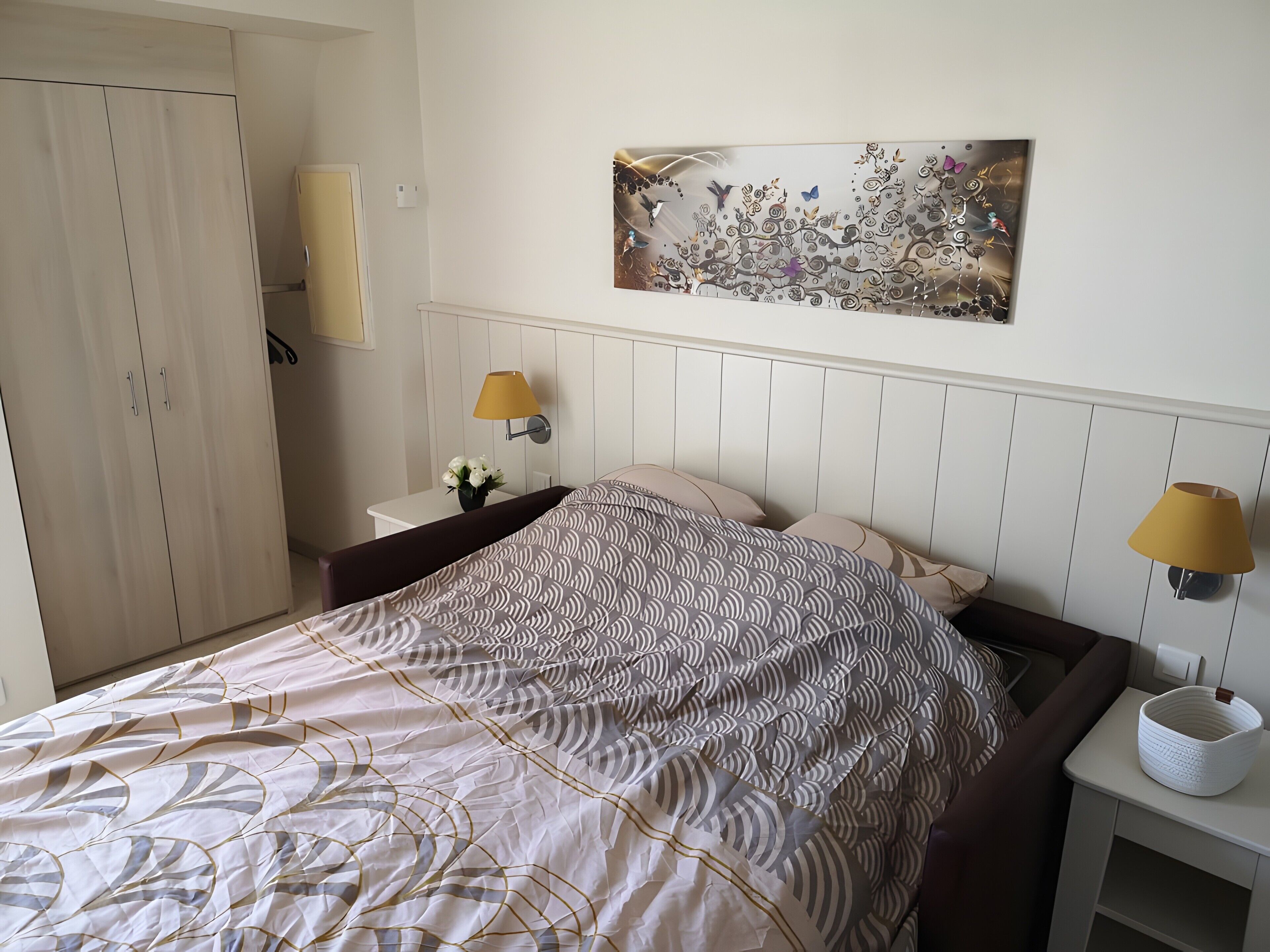 Une chambre à coucher comprenant un lit, deux lampes de chevet, une œuvre d’art accrochée au mur, une armoire et une table de chevet ornée d’un vase de fleurs.