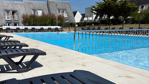 A swimming pool with lounge chairs and a building with a sloped roof in the background. - La Perle de l'Océan – Pool and Terrace (Guidel)