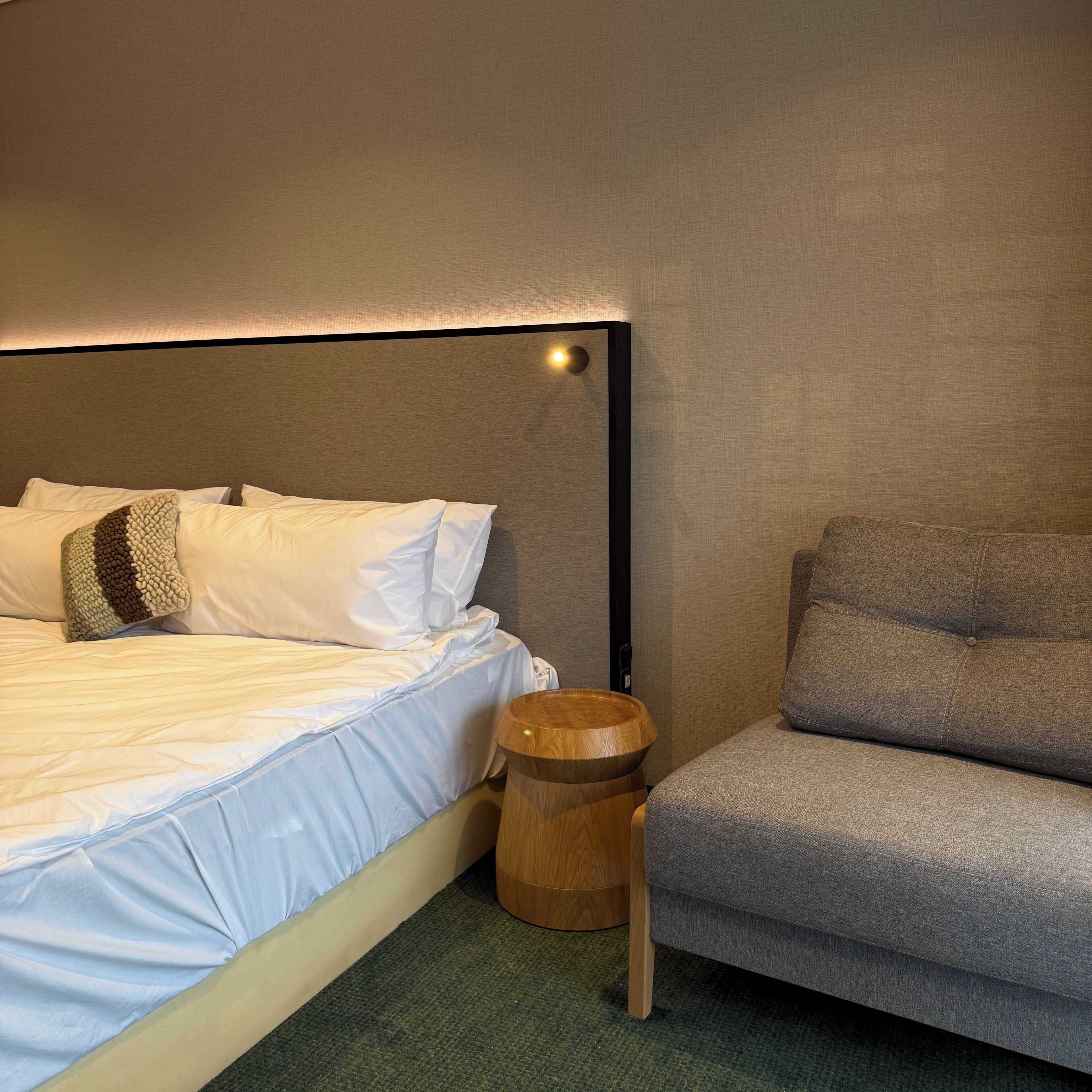 A neatly made bed with white linens and a grey upholstered chair beside it.