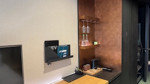 Une chambre d’hôtel avec un bureau en bois, une télécommande noire, une machine à café et une étagère contenant des verres à vin et un récipient.