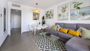 Ein modernes Wohnzimmer mit grauem Sofa, weißen Stühlen, einem Glastisch und tropisch gestalteter Wandkunst.