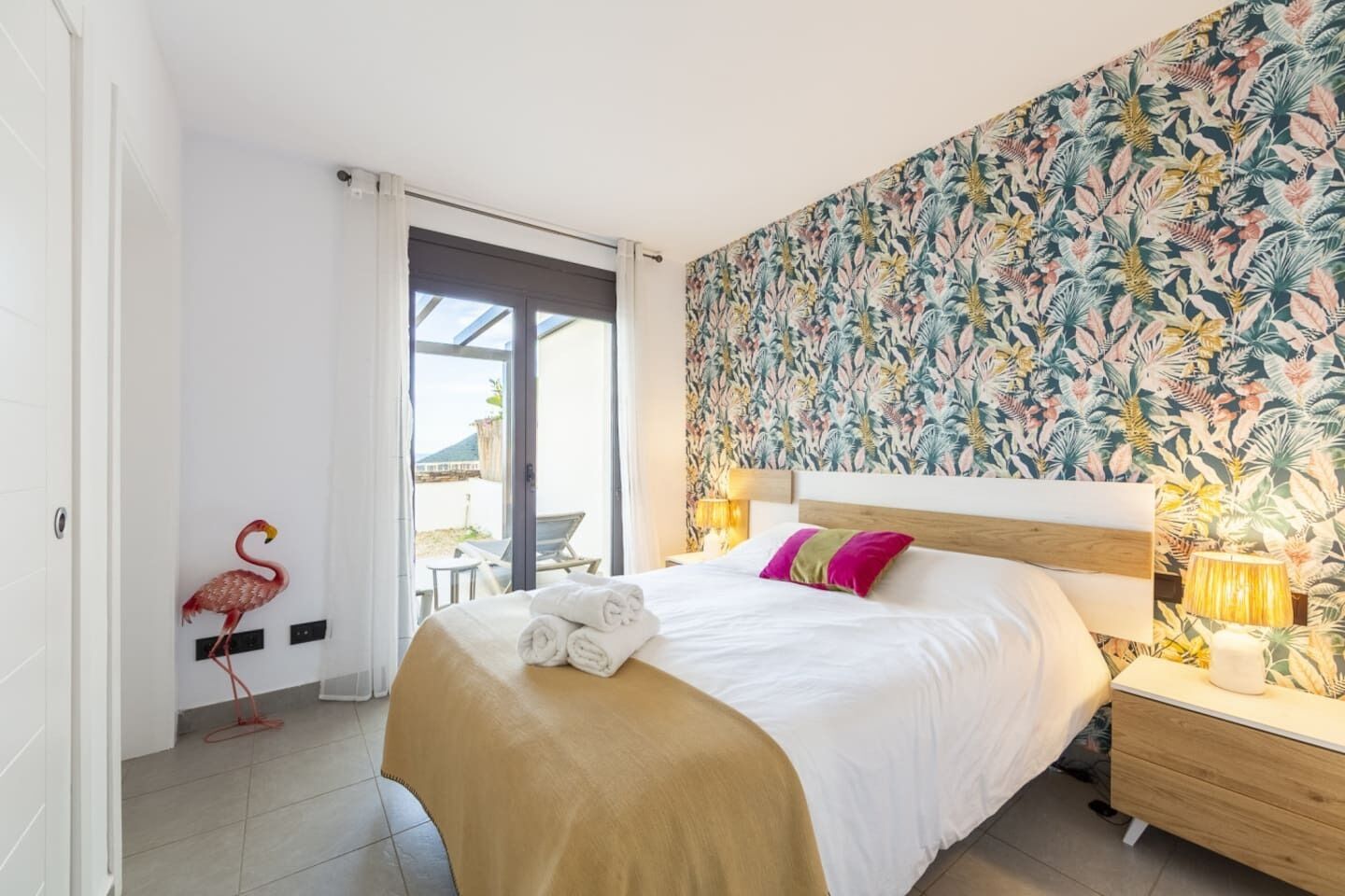 A bedroom with a bed, a nightstand, a lamp, a wall with a tropical pattern, a door, and a flamingo decoration.
