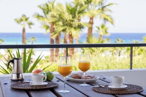A table with a coffee pot, two glasses of orange juice, and a plate of pastries on a balcony overlooking the ocean.