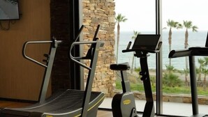 A gym with treadmills and exercise equipment, a stone wall, and a large window with a view of palm trees and the ocean. - Amazing Sea views # Macenas Resort # 2BR # Sunbeds (Mojácar)