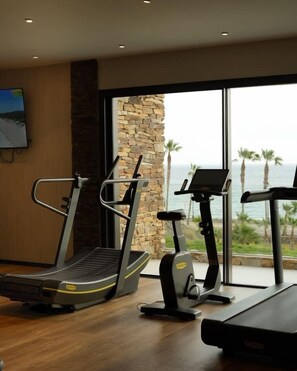 A gym with treadmills and exercise equipment, a stone wall, and a large window with a view of palm trees and the ocean.