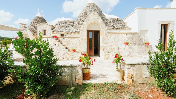 Ein traditionelles Steinhaus mit Kegeldach, einer Holztür und roten Blumen in Töpfen im Eingangsbereich.