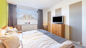 A bedroom with a bed, a television on a wooden cabinet, a window with curtains, and a radiator.