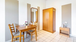 A room with a wooden dining table, two chairs, a mirror, a wardrobe, and a small bedside table with a flower vase.