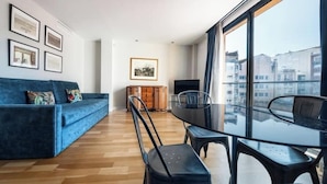 Een moderne woonkamer met een blauwe bank, een glazen eettafel en stoelen. Op de muur hangen ingelijste foto’s en er staat een televisie op de achtergrond.