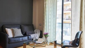 Un saló modern amb un sofà de color fosc, una taula de centre de vidre i una vista d'un edifici a través de grans finestres.