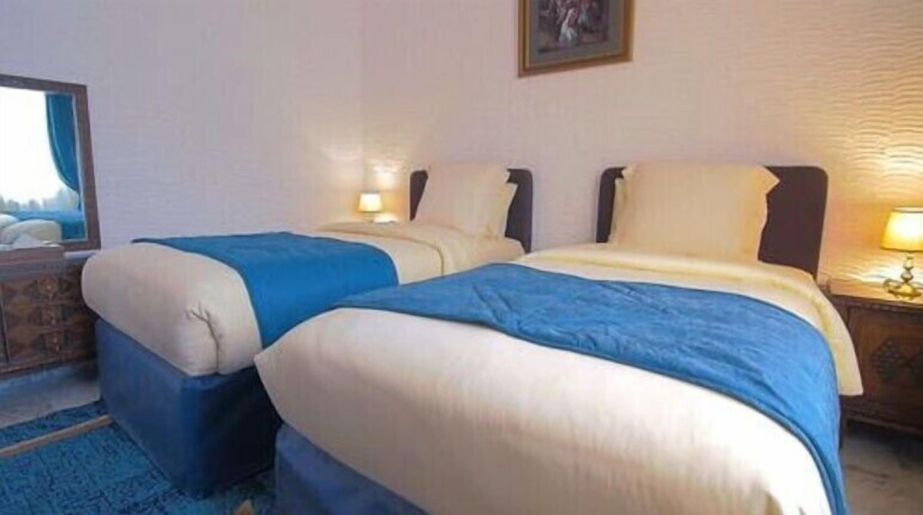 A hotel room with two beds, each with a blue and white duvet, a bedside table with a lamp, and a mirror on the wall.