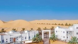 Un complexe hôtelier situé dans le désert, avec des bâtiments blancs, des palmiers et des dunes de sable en arrière-plan.