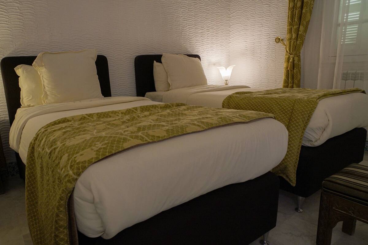 Ett hotellrum med två sängar, båda med vita sängkläder och gröna, mönstrade plädar. Det finns sänglampor på träbord som står vid sängarna.