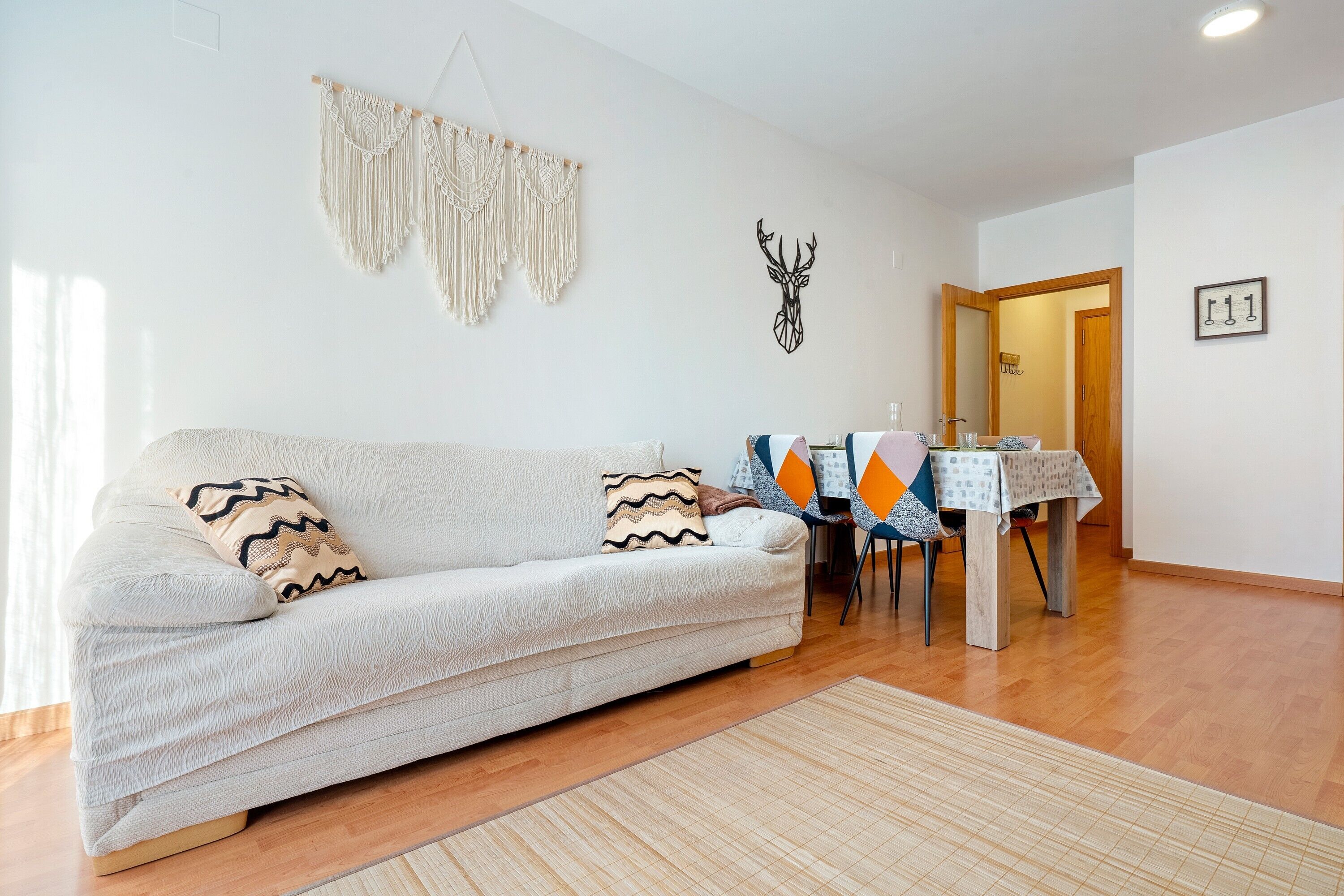 Een woonkamer met een bank, een macramé wanddecoratie, een herten muurstickers en een eethoek met een tafel en stoelen.