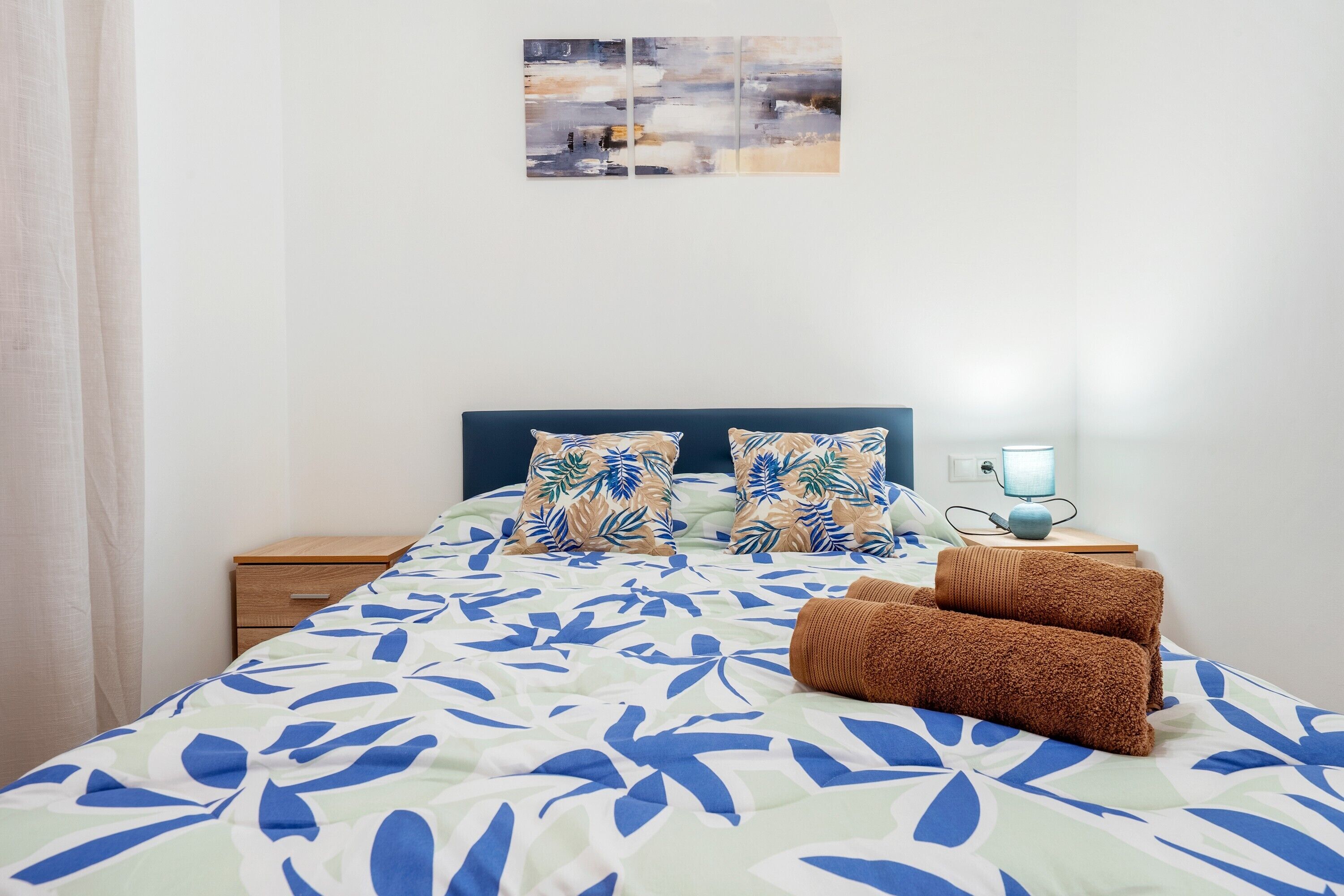 Un letto con copripiumino a motivo di foglie, due cuscini dal design tropicale, una lampada da comodino e un piccolo comodino in legno.